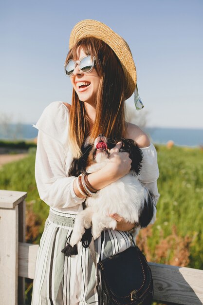 Feliz joven elegante en campo, sosteniendo un perro, feliz estado de ánimo positivo, verano, sombrero de paja, traje de estilo bohemio, gafas de sol, sonriendo