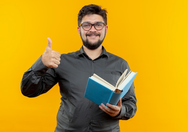 Feliz joven empresario con gafas sosteniendo el libro con el pulgar hacia arriba aislado sobre fondo amarillo