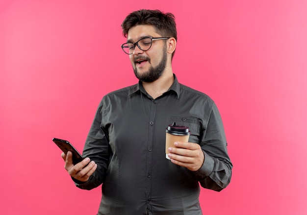 Feliz joven empresario con gafas sosteniendo una taza de café y mirando el teléfono en la mano