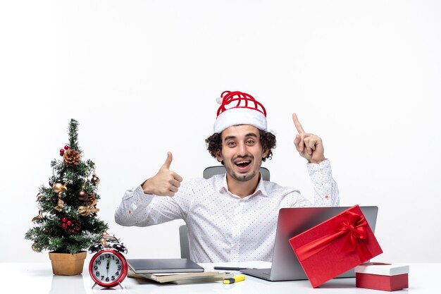 Feliz joven empresario positivo con divertido sombrero de santa claus apuntando hacia arriba y haciendo un gesto de ok en la oficina sobre fondo blanco.