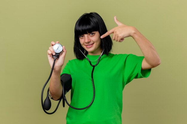 Feliz joven enferma con estetoscopio mostrando esfigmomanómetro apuntando hacia él mirando al frente aislado en la pared verde oliva con espacio de copia