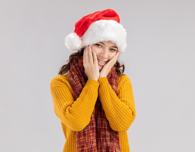 Feliz joven eslava con gorro de Papá Noel y bufanda alrededor del cuello pone las manos en la cara