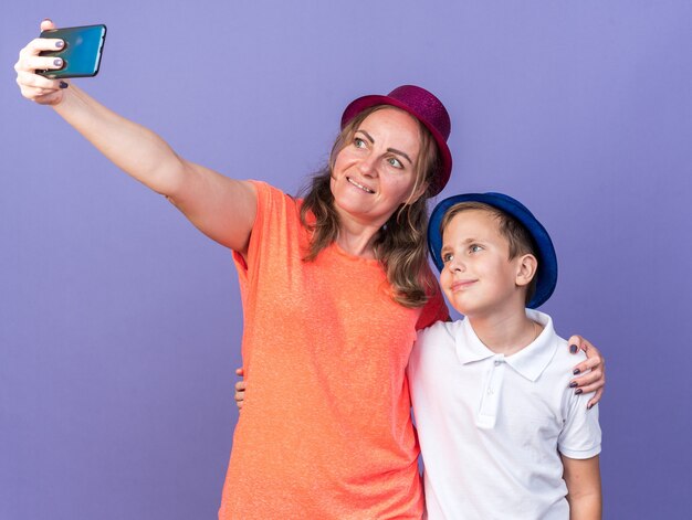 Feliz joven eslavo con gorro de fiesta azul tomando selfie con su madre vistiendo gorro de fiesta violeta aislado en la pared púrpura con espacio de copia