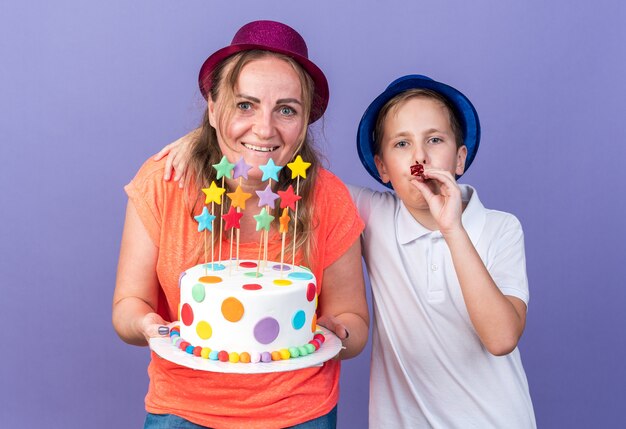 Feliz joven eslavo con sombrero de fiesta azul soplando silbato de fiesta de pie con su madre con sombrero de fiesta violeta sosteniendo pastel de cumpleaños aislado en la pared púrpura con espacio de copia