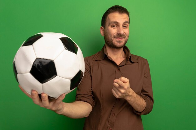 Feliz joven estirando la pelota de fútbol hacia el frente mirando a la cámara apuntando a la pelota aislada en la pared verde