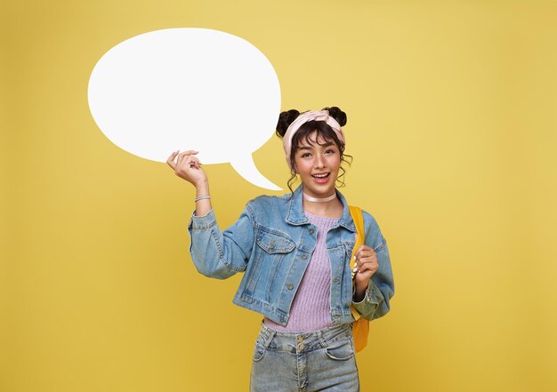 Feliz joven estudiante asiática sosteniendo un bocadillo de diálogo vacío aislado sobre un fondo amarillo