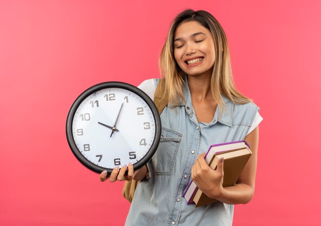 Feliz joven estudiante bonita vestida con bolsa trasera sosteniendo libros y reloj con los ojos cerrados aislados en rosa