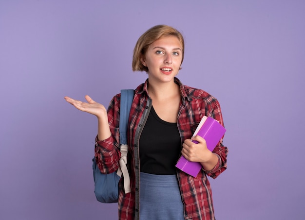 Feliz joven estudiante eslava con mochila sostiene el libro y el cuaderno mantiene la mano abierta