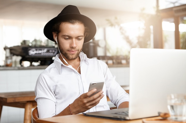 Feliz joven estudiante con sombrero elegante y auriculares escuchando música en su teléfono móvil, mientras se relaja en el interior en la acogedora cafetería, sentado a la mesa con la computadora portátil.