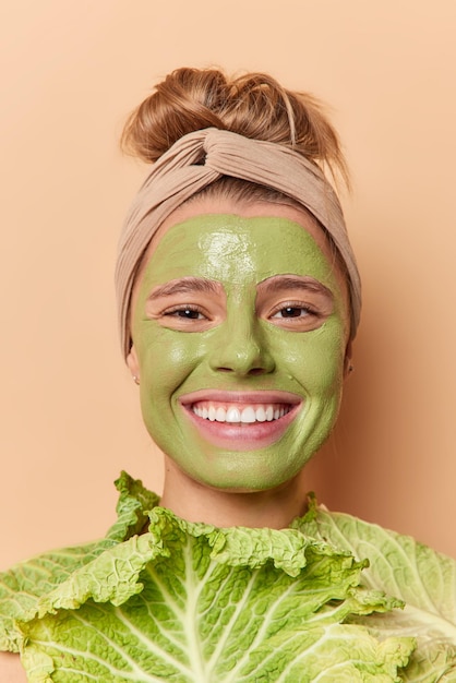 Foto gratuita feliz joven europea sostiene repollo fresco usa máscara de belleza verde sonríe con dientes muestra dientes blancos usa diadema aislada sobre fondo beige tratamientos faciales y concepto de bienestar