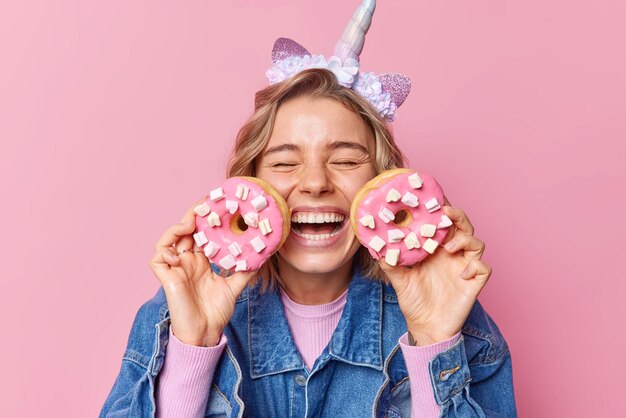 Feliz joven europea vestida con una chaqueta de mezclilla de moda sostiene dos deliciosos donuts con mashmallow cerca de la cara tiene un diente dulce que se ríe felizmente mantiene los ojos cerrados aislados sobre una pared rosa