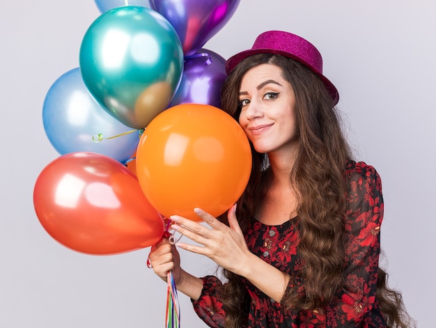 Feliz joven fiestera con sombrero de fiesta sosteniendo y tocando globos mirando a la cámara aislada en la pared blanca