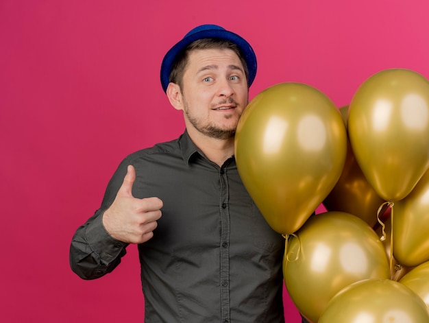 Feliz joven fiestero con sombrero azul de pie junto a globos mostrando el pulgar hacia arriba aislado en rosa