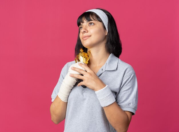 Feliz joven fitness chica con diadema con mano vendada sosteniendo el trofeo mirando hacia arriba sintiéndose agradecido de pie sobre fondo rosa