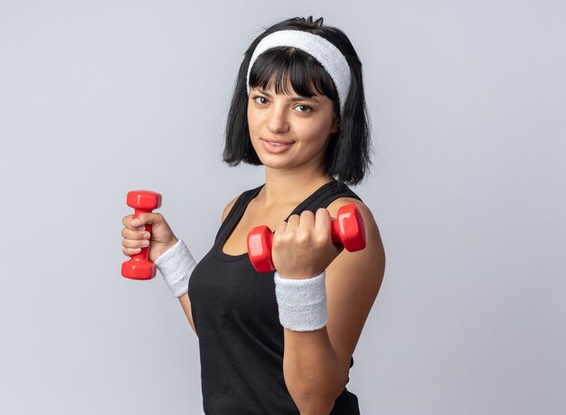 Feliz joven fitness chica con diadema sosteniendo pesas haciendo ejercicios mirando confiados de pie sobre fondo blanco.