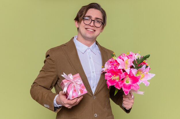 Feliz joven con gafas sosteniendo presente y ramo de flores sonriendo alegremente yendo a felicitar con el día internacional de la mujer de pie sobre la pared verde