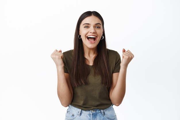 Feliz joven ganando el premio, haciendo bomba de puño y sonriendo emocionado, triunfando como meta, celebrando la victoria, gritando asombrado, pared blanca