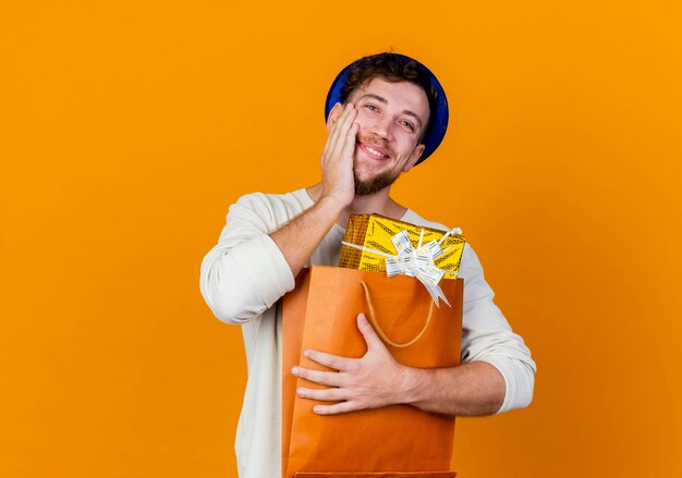 Feliz joven guapo chico partido eslavo con sombrero de fiesta sosteniendo cajas de regalo en una bolsa de papel manteniendo la mano en la cara aislada sobre fondo naranja con espacio de copia