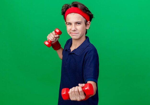 Feliz joven guapo deportivo con diadema y muñequeras con aparatos dentales de pie en la vista de perfil sosteniendo y estirando pesas aisladas sobre fondo verde con espacio de copia