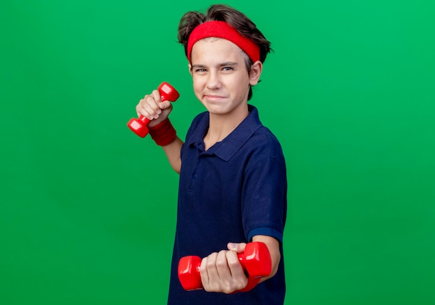 Feliz joven guapo deportivo con diadema y muñequeras con aparatos dentales de pie en la vista de perfil sosteniendo y estirando pesas aisladas sobre fondo verde con espacio de copia