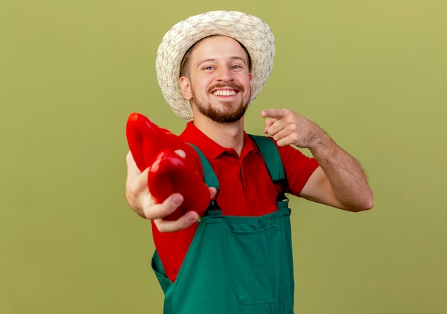 Feliz joven guapo jardinero eslavo en uniforme y sombrero mirando y señalando estirando pimientos aislados en la pared verde oliva con espacio de copia