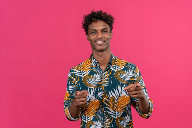 Feliz joven guapo de piel oscura con cabello rizado en hojas camisa estampada apuntando a la cámara con los dedos índices