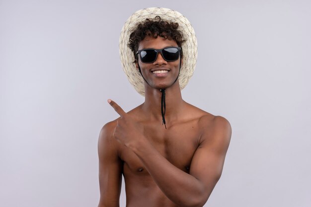 Un feliz joven guapo de piel oscura con cabello rizado con sombrero para el sol y gafas de sol apuntando con el dedo índice mientras