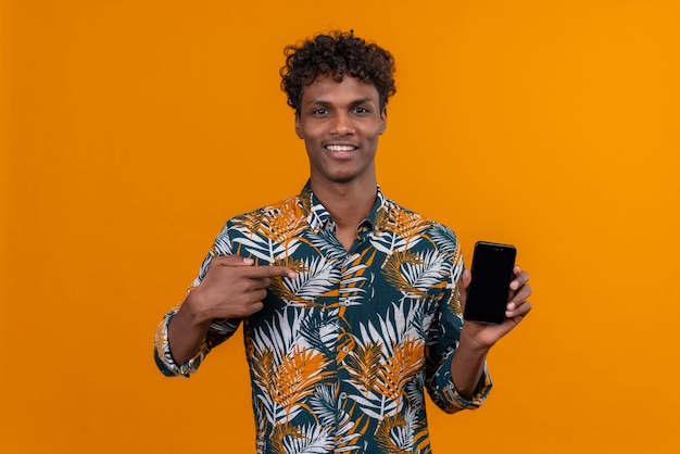 Feliz joven guapo de piel oscura con pelo rizado apuntando al teléfono inteligente en blanco con el dedo índice