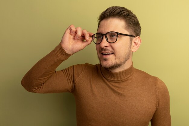 Feliz joven guapo rubio vistiendo y agarrando gafas mirando al lado aislado en la pared verde oliva