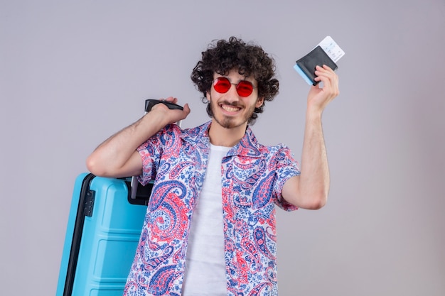 Feliz joven guapo viajero rizado hombre con gafas de sol sosteniendo billetera y boletos de avión con maleta en espacio en blanco aislado con espacio de copia