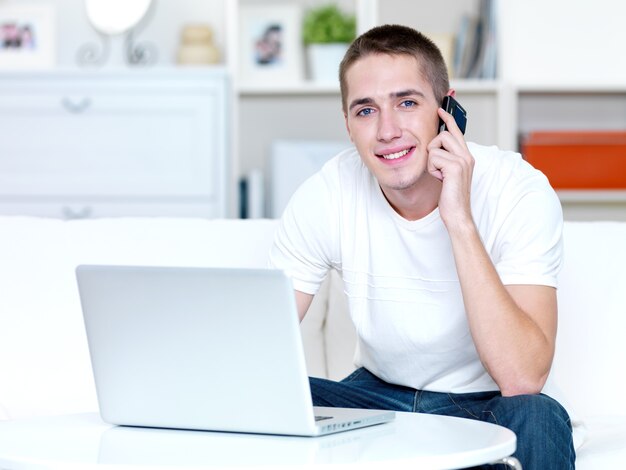 Feliz joven habla por teléfono y trabaja en la computadora portátil en casa