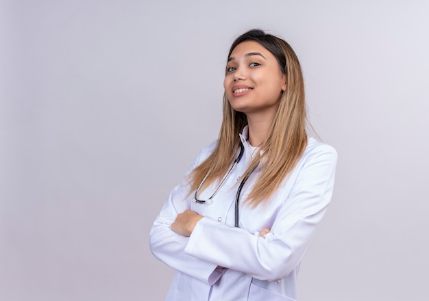 Feliz joven hermosa médico vistiendo bata blanca con estetoscopio mirando confiado de pie con los brazos cruzados.