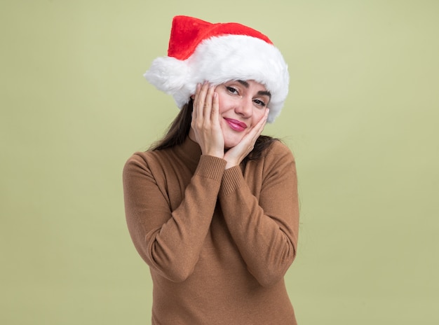 Foto gratuita feliz joven hermosa niña con sombrero de navidad cubierto las mejillas con las manos aisladas en la pared verde oliva