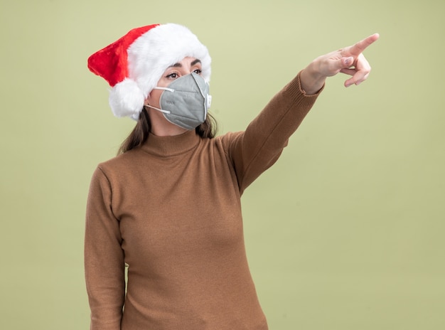 Foto gratuita feliz joven hermosa niña con sombrero de navidad con puntos de máscara médica en el lado aislado sobre fondo verde oliva