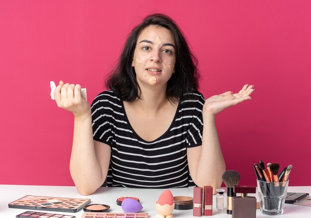 Feliz joven hermosa se sienta a la mesa con herramientas de maquillaje aplicando crema tonificante extendiendo las manos aisladas en la pared rosa