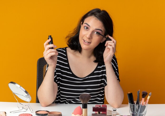 Feliz joven hermosa se sienta a la mesa con herramientas de maquillaje aplicando rímel aislado en la pared naranja