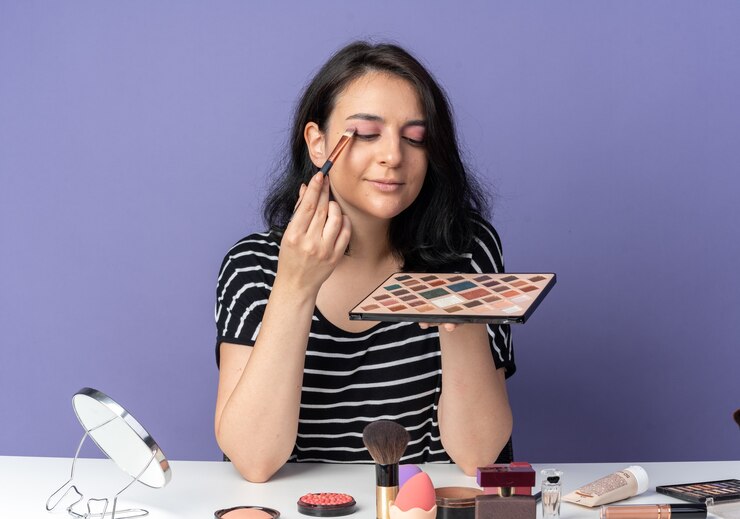 Foto gratuito feliz joven hermosa se sienta a la mesa con herramientas de maquillaje aplicando sombra de ojos con pincel de maquillaje aislado en la pared azul