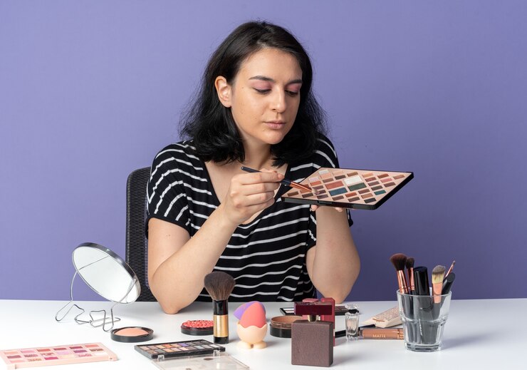 Foto gratuito feliz joven hermosa se sienta a la mesa con herramientas de maquillaje aplicando sombra de ojos con pincel de maquillaje aislado en la pared azul