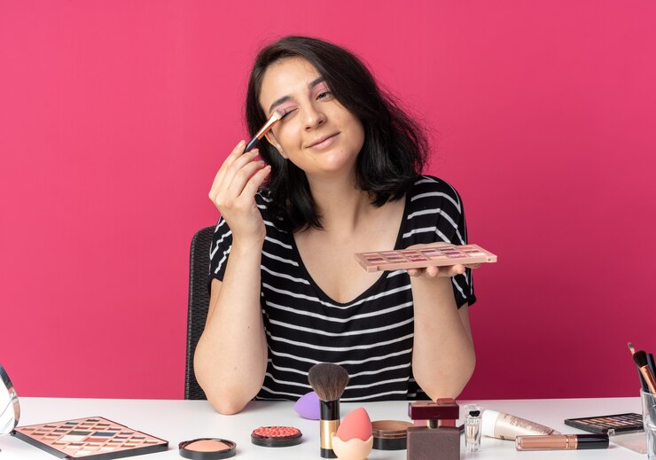 Foto gratuito feliz joven hermosa se sienta a la mesa con herramientas de maquillaje aplicando sombra de ojos con pincel de maquillaje aislado en la pared rosa