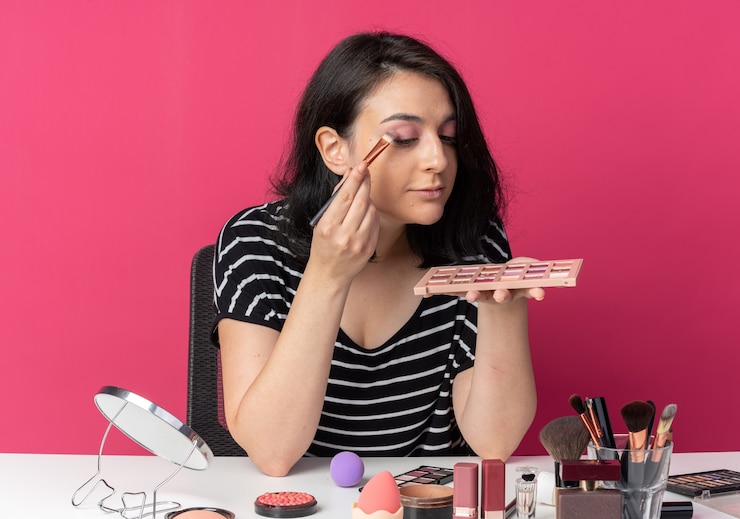 Foto gratuito feliz joven hermosa se sienta a la mesa con herramientas de maquillaje aplicando sombra de ojos con pincel de maquillaje aislado en la pared rosa