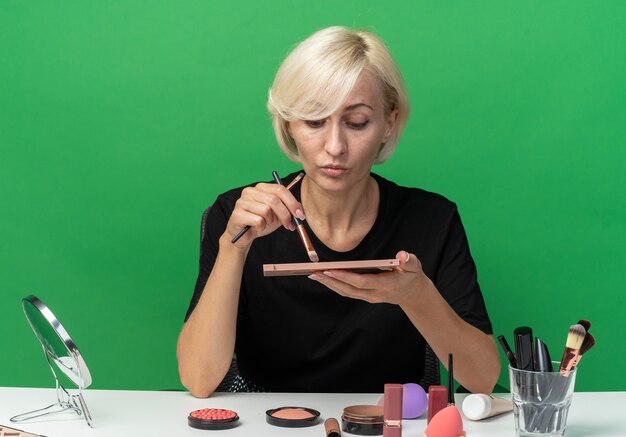 Feliz joven hermosa se sienta a la mesa con herramientas de maquillaje aplicando sombra de ojos con pincel de maquillaje aislado en la pared verde