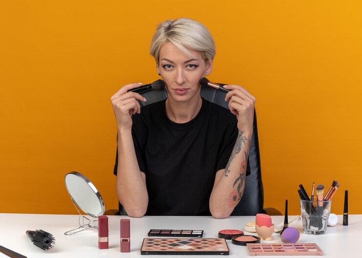 Foto gratuito feliz joven hermosa se sienta a la mesa con herramientas de maquillaje con pinceles en polvo en la cara aislada en la pared naranja