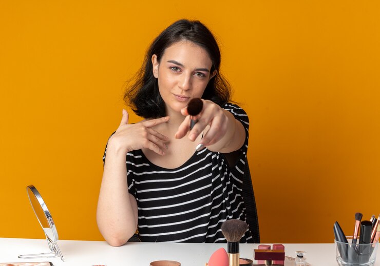 Foto gratuito feliz joven hermosa se sienta a la mesa con herramientas de maquillaje sosteniendo un cepillo de polvo aislado en la pared naranja