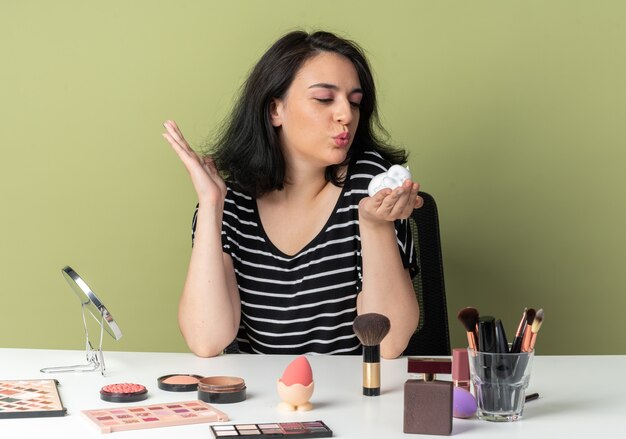 Feliz joven hermosa se sienta a la mesa con herramientas de maquillaje sosteniendo y mirando crema para el cabello aislado en la pared verde oliva