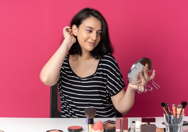 Feliz joven hermosa se sienta a la mesa con herramientas de maquillaje sosteniendo y mirando el espejo poniendo la mano en la cabeza aislada en la pared rosa