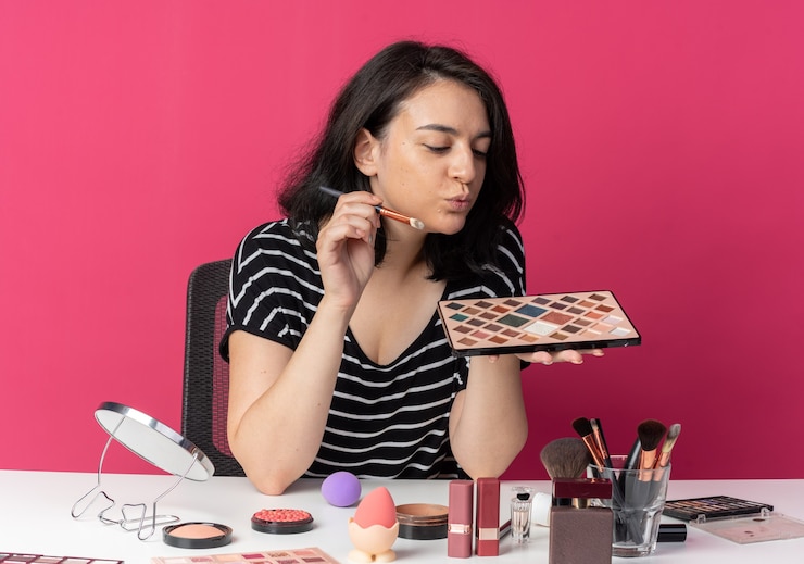 Foto gratuito feliz joven hermosa se sienta a la mesa con herramientas de maquillaje sosteniendo y mirando la paleta de sombras de ojos con pincel de maquillaje aislado en la pared rosa