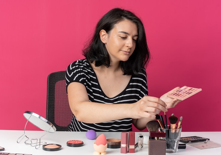 Foto gratuito feliz joven hermosa se sienta a la mesa con herramientas de maquillaje sosteniendo la paleta de sombra de ojos con pincel de maquillaje aislado en la pared rosa