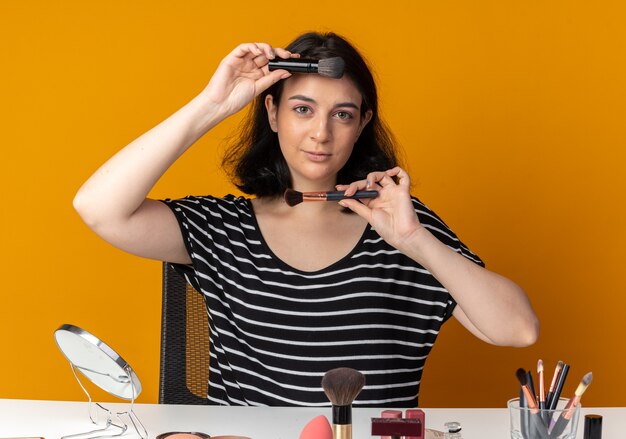 Feliz joven hermosa se sienta a la mesa con herramientas de maquillaje sosteniendo pinceles de maquillaje en la frente y el mentón aislado en la pared naranja