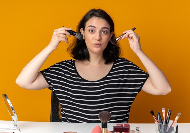 Feliz joven hermosa se sienta a la mesa con herramientas de maquillaje sosteniendo pinceles en polvo alrededor de la cara aislada en la pared naranja