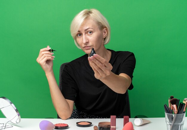 Feliz joven hermosa se sienta a la mesa con herramientas de maquillaje sosteniendo rímel en cámara aislada en pared verde